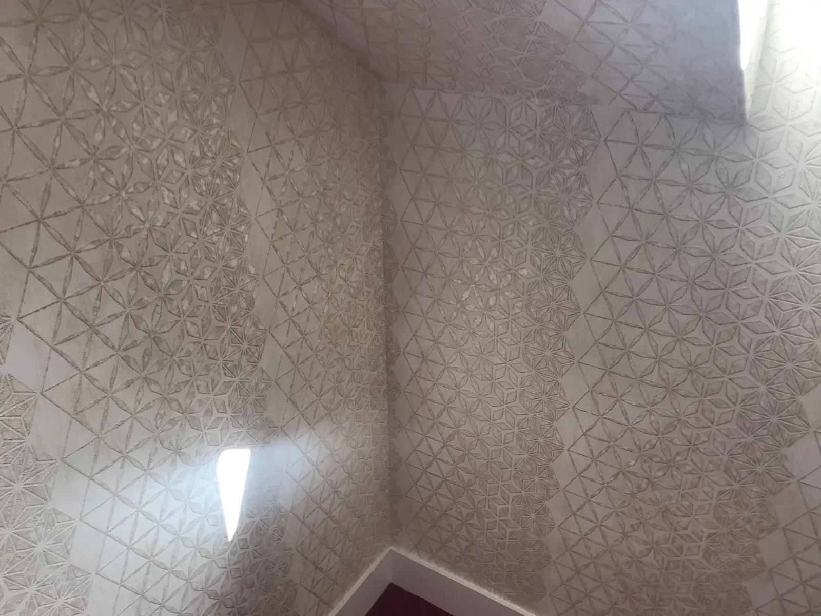Papier peint beige clair à motif géométrique en relief, pose soignée avec raccords parfaits, texture mise en valeur par la lumière.