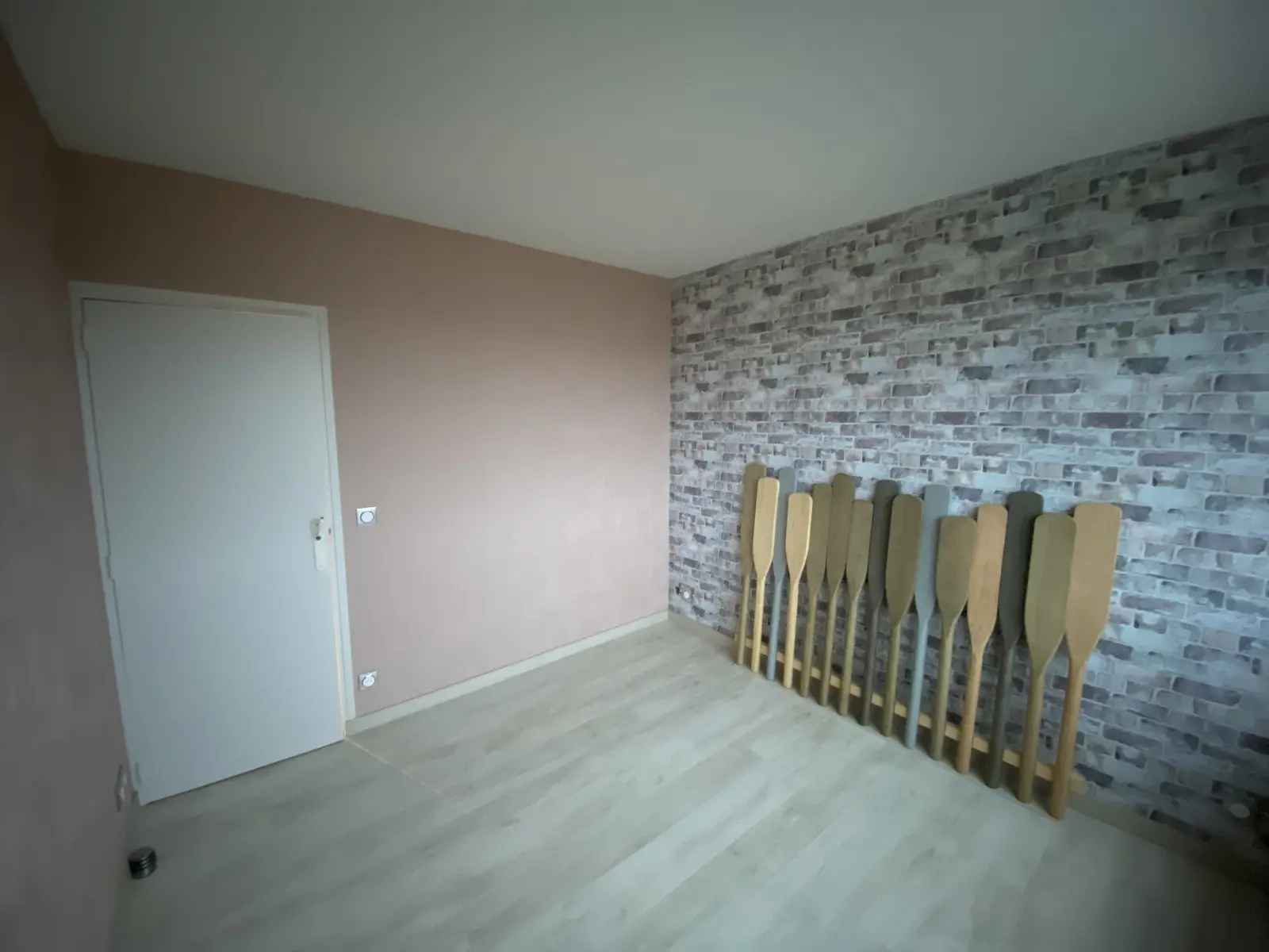 Vue d'angle sur une chambre rénovée, mettant en valeur le contraste entre le mur en papier peint imitation brique gris clair et les murs rose pâle. Ambiance moderne et apaisante.