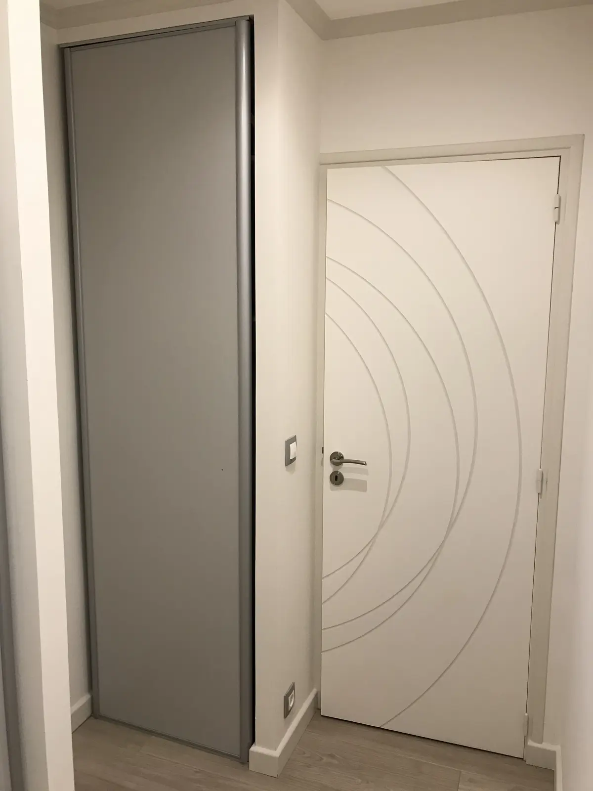 Couloir rénové avec porte blanche moderne : lignes courbes en relief aux rainures soulignées d'une teinte foncée. Plinthes de plafond et encadrement assortis, créant une harmonie élégante et contemporaine.
