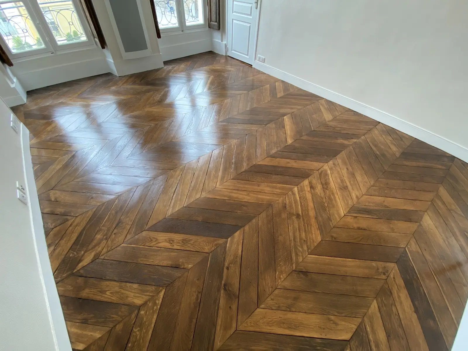 Sol en parquet chevron, récemment vitrifié. Le parquet, de couleur brun foncé, présente un aspect brillant et lisse témoignant d'une finition soignée. La disposition en chevrons est régulière et nette.